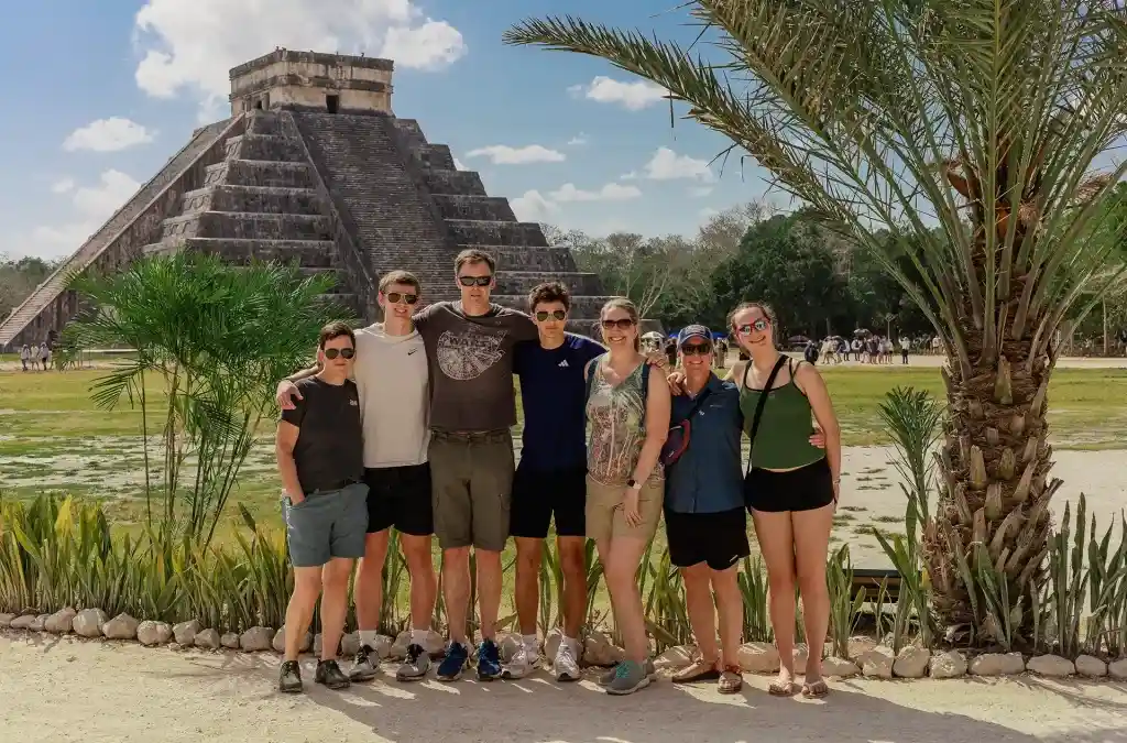 Smiling family group posing in front of the Kukulcan Pyramid at Chichen Itza on a sunny day.