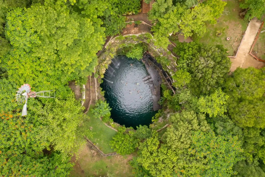Aerial view of an open cenote in the Yucatan peninsula, showcasing its clear blue water surrounded by dense, green jungle.
