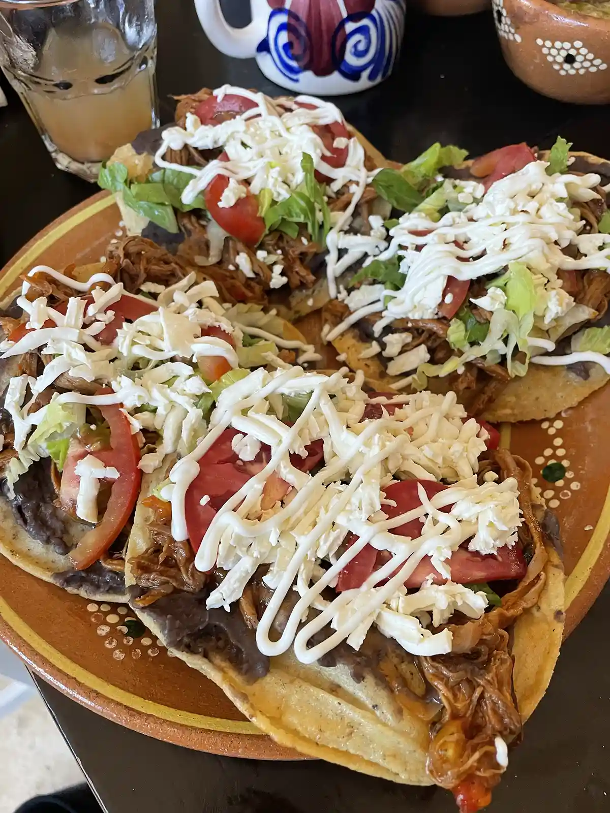 A plate with four tostadas topped with shredded chicken, refried beans, lettuce, tomato slices, shredded cheese, and drizzled with sour cream—perfect for anyone seeking low budget food in Tulum. 