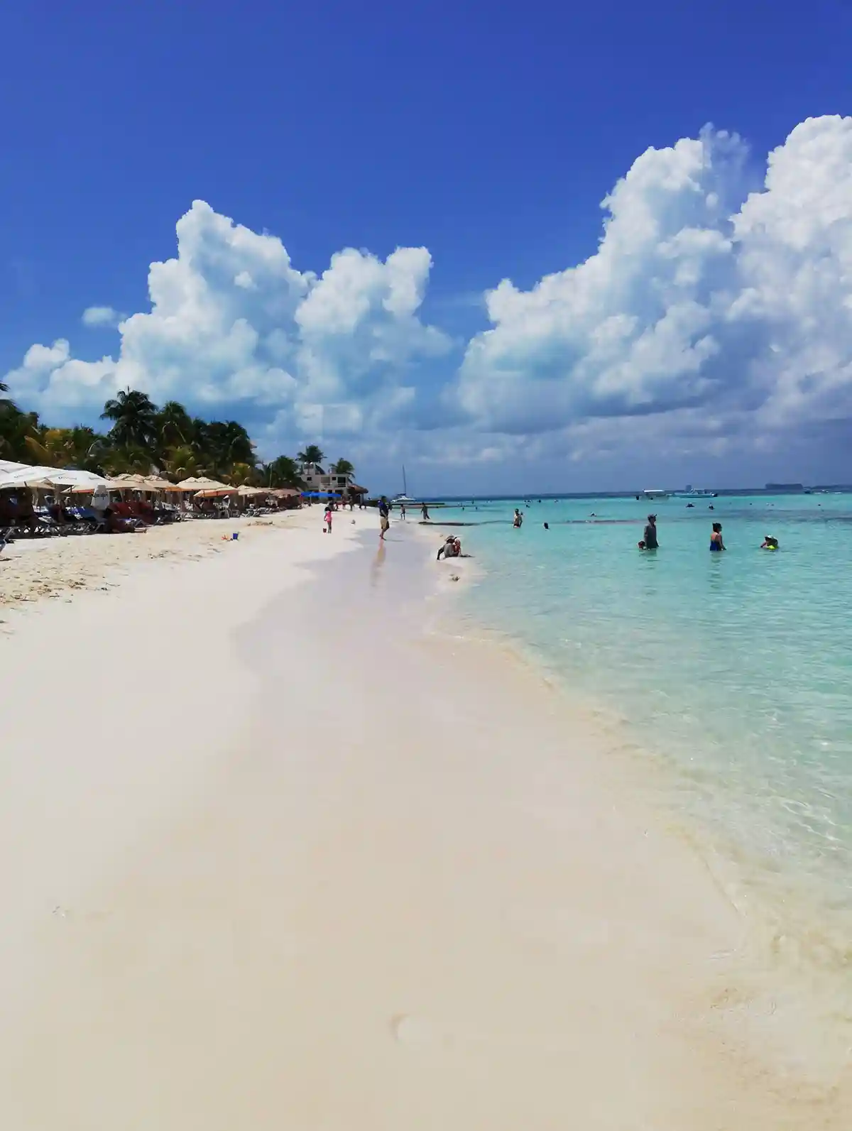 Tourists enjoying a sunny day at a free public beach in Tulum, with clear turquoise water, white sand, and umbrellas lining the shore.