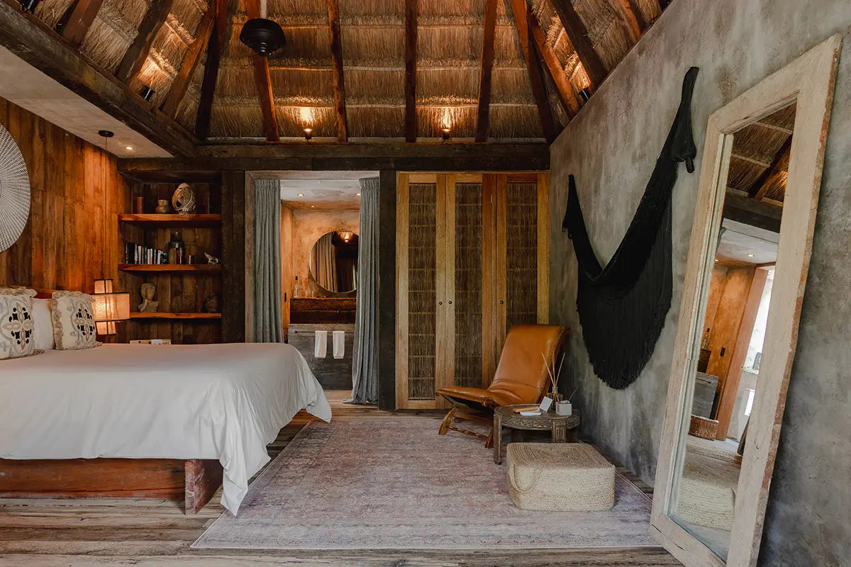 Rustic, eco-friendly Tulum hotel beach house bedroom with wooden furniture, leather chair, and sea view.