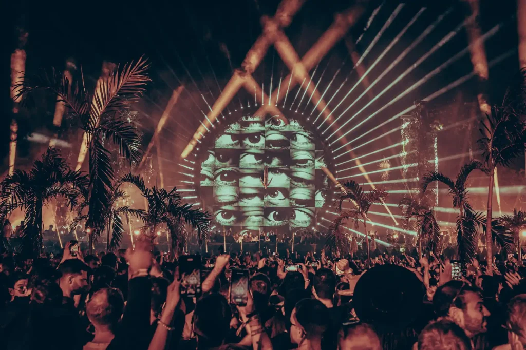 A vibrant night at Zamna Festival Tulum with a large crowd gathered under palm trees, featuring a dramatic circular stage with a visual display of eyes and radiant light beams creating a mesmerizing atmosphere.