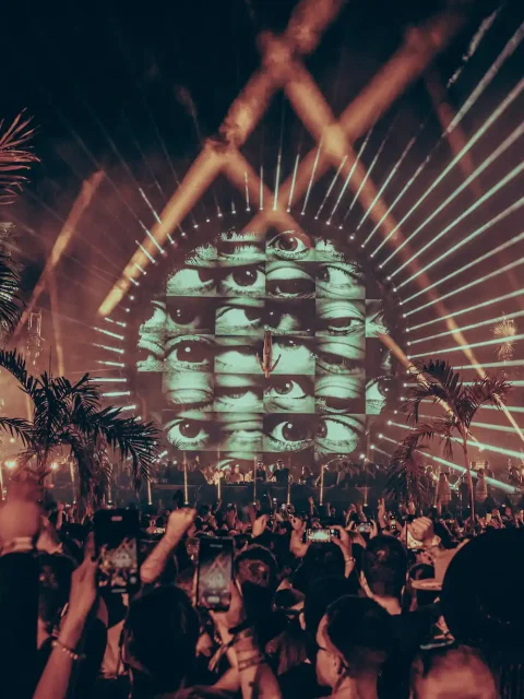 A vibrant night at Zamna Festival Tulum with a large crowd gathered under palm trees, featuring a dramatic circular stage with a visual display of eyes and radiant light beams creating a mesmerizing atmosphere.