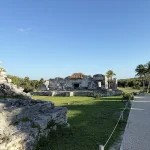 “Ancient Mayan ruins at the Tulum Archaeological Site, surrounded by greenery and a clear blue sky, with a stone pathway leading through.