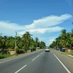 A car drives along a tropical highway lined with palm trees under a bright blue sky.