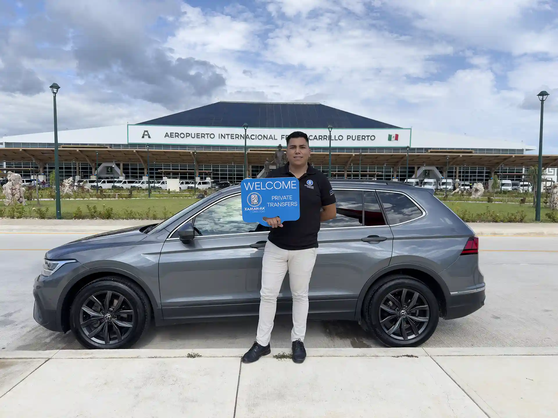 Private transfer driver holding welcome sign in front of Tulum Airport and a gray SUV.