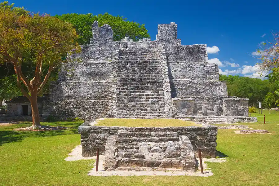 The main pyramid at the El Meco Mayan ruins in Cancún, featuring a stone staircase leading to the top, is surrounded by manicured grass and trees.
