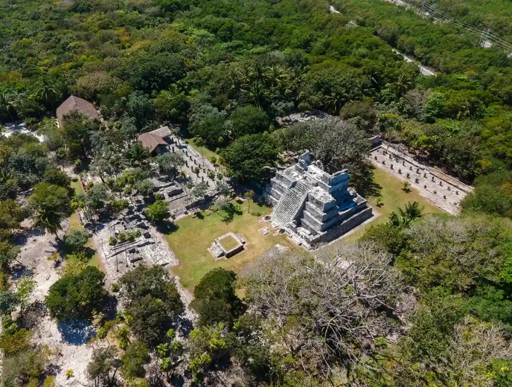 Aerial view of El Meco Mayan ruins near Cancún, featuring a central stepped pyramid surrounded by lush jungle and smaller stone structures.