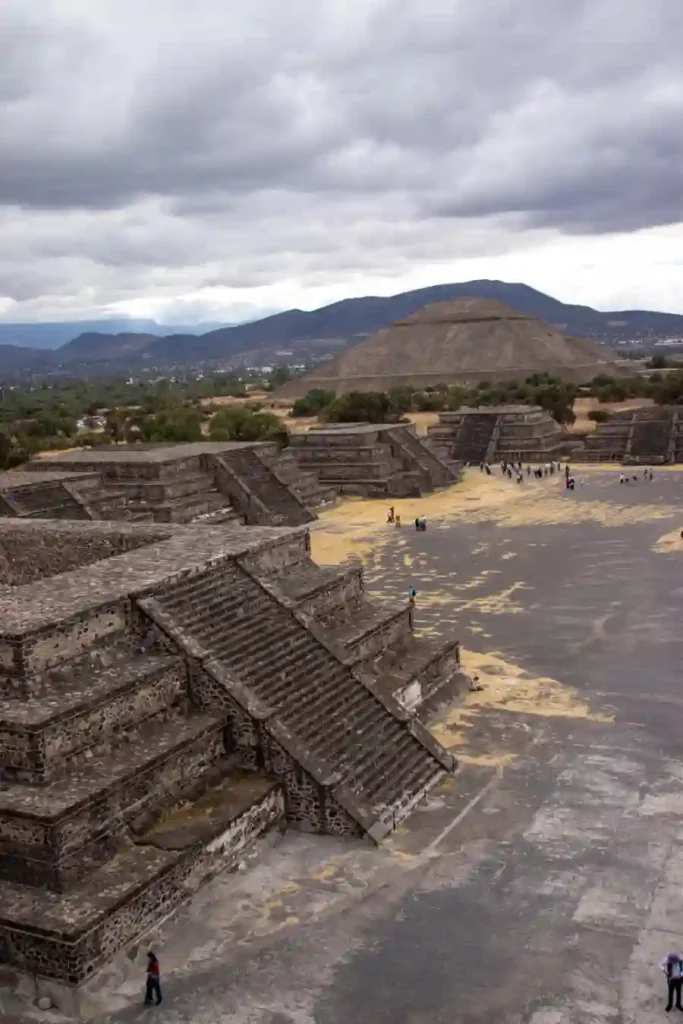 A panoramic view of the stepped stone pyramids at Teotihuacán under a cloudy sky, with tourists walking along the ancient avenue.