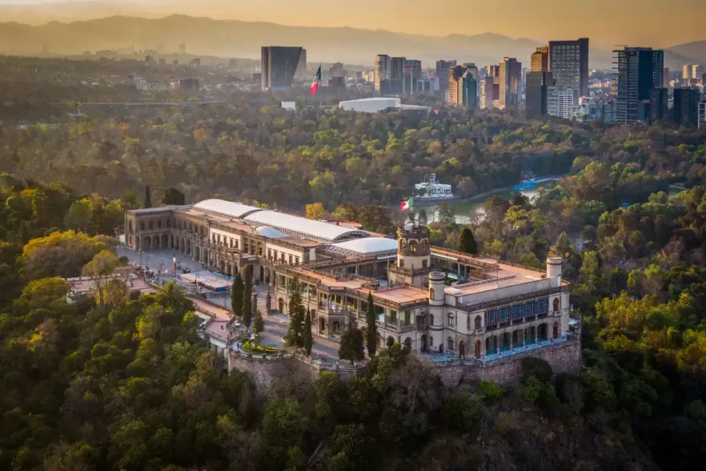 The Chapultepec Castle surrounded by lush forest and the skyline of Mexico City in the background during sunset.