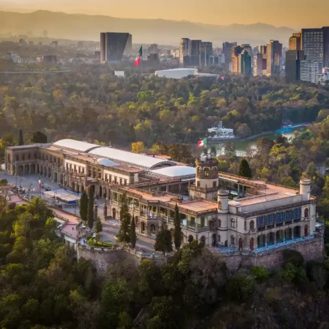 The Chapultepec Castle surrounded by lush forest and the skyline of Mexico City in the background during sunset.