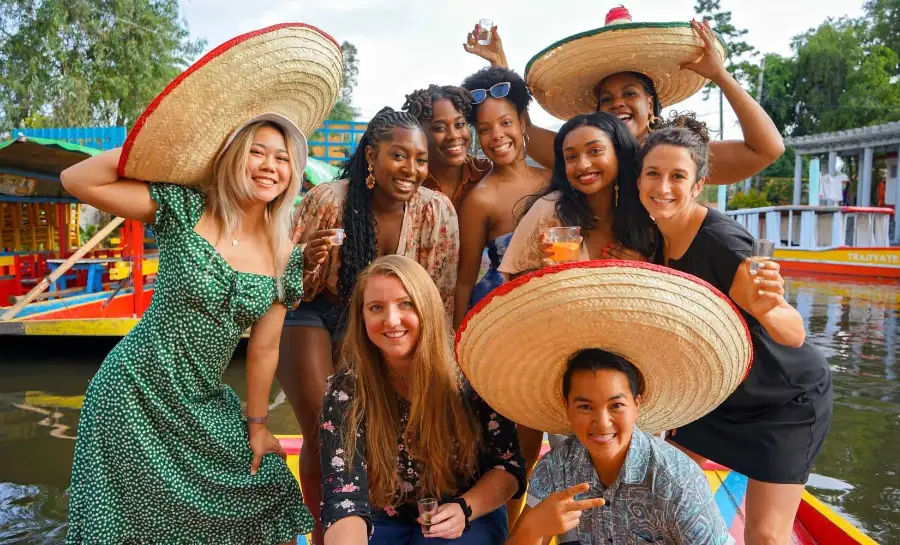 Group of friends wearing sombreros and celebrating with drinks on a colorful trajinera boat in the canals of Xochimilco, Mexico City.