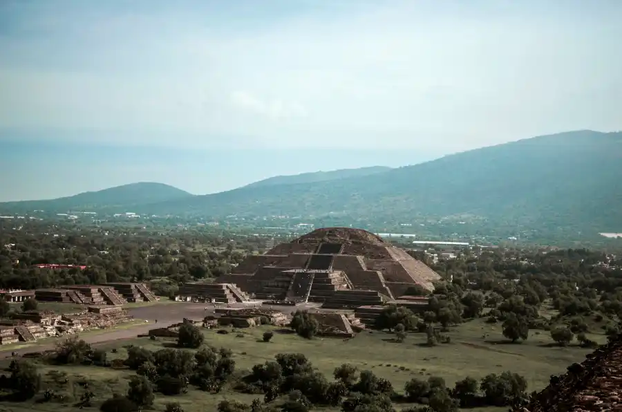 A distant aerial view of the Pyramid of the Moon in Teotihuacán, nestled in a green valley under a soft blue sky.