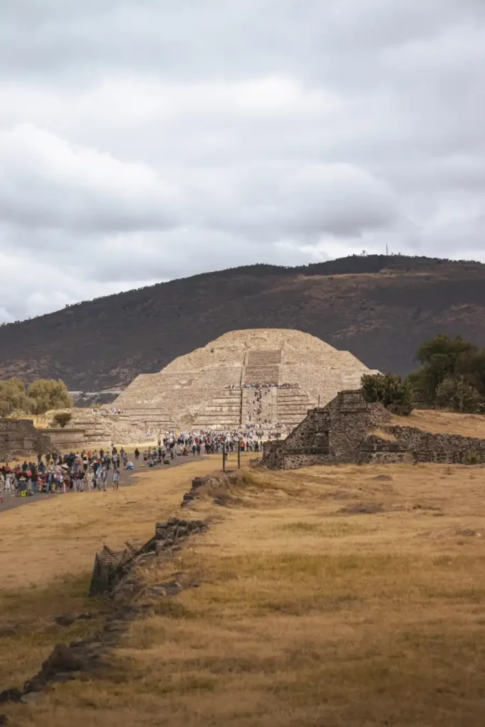 A wide-angle photo showing a crowd of visitors walking toward the Pyramid of the Moon, framed by a dry, golden landscape.