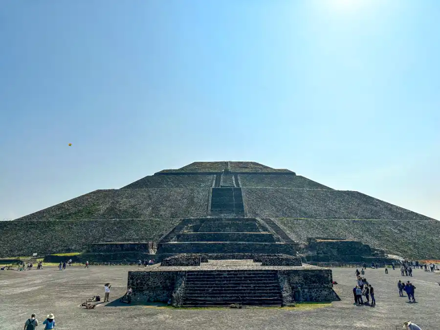 A wide, sunlit view of the Pyramid of the Sun, with scattered visitors walking around the base under a clear sky.