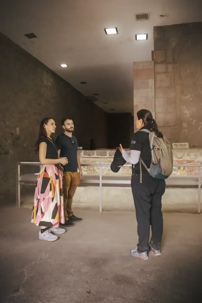 A private tour guide explaining mural art to two visitors inside an ancient stone-walled exhibition room in Teotihuacán.