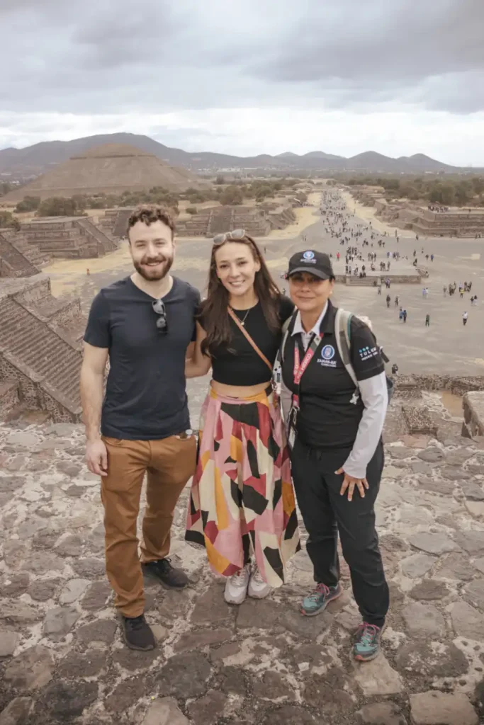 Couple with local guide standing atop the Pyramid of the Moon in Teotihuacan, Mexico, with the Pyramid of the Sun and Avenue of the Dead in the background.