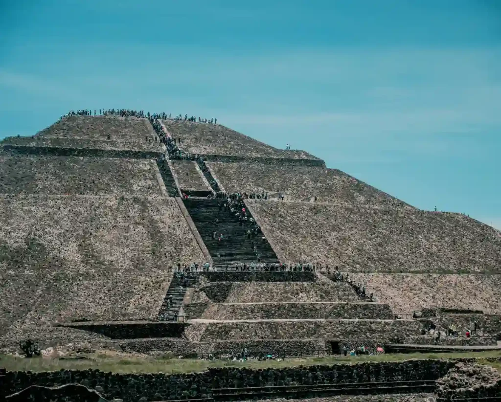 A front-facing shot of the Pyramid of the Sun with many tourists ascending the central staircase under a bright blue sky.
