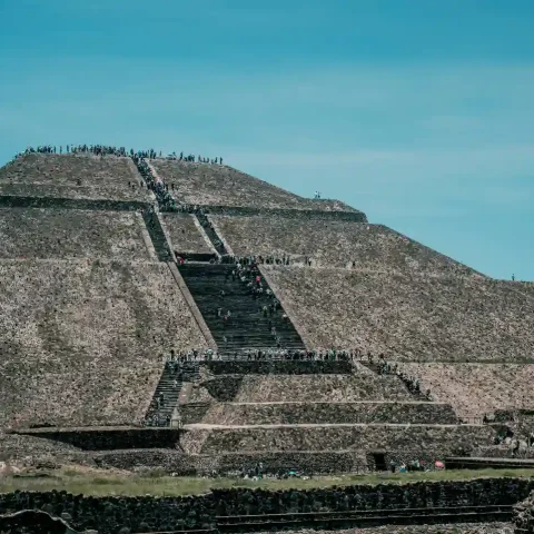 A front-facing shot of the Pyramid of the Sun with many tourists ascending the central staircase under a bright blue sky.