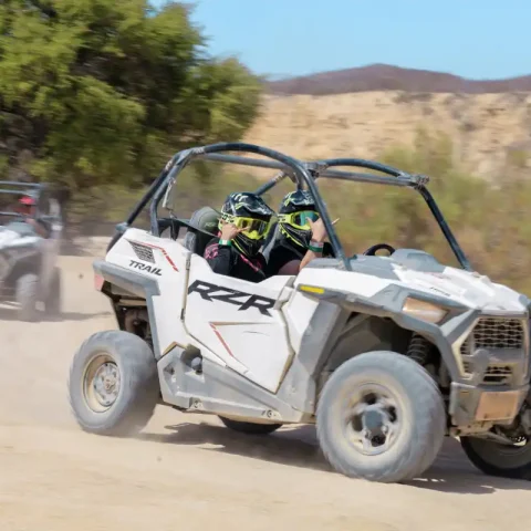 Two riders driving a UTV through desert trails in Cabo San Lucas with dust clouds and rugged terrain.