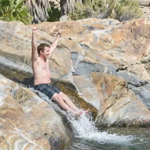 Man sliding down natural rock waterslide with arms raised at Fox Canyon in Cabo San Lucas.