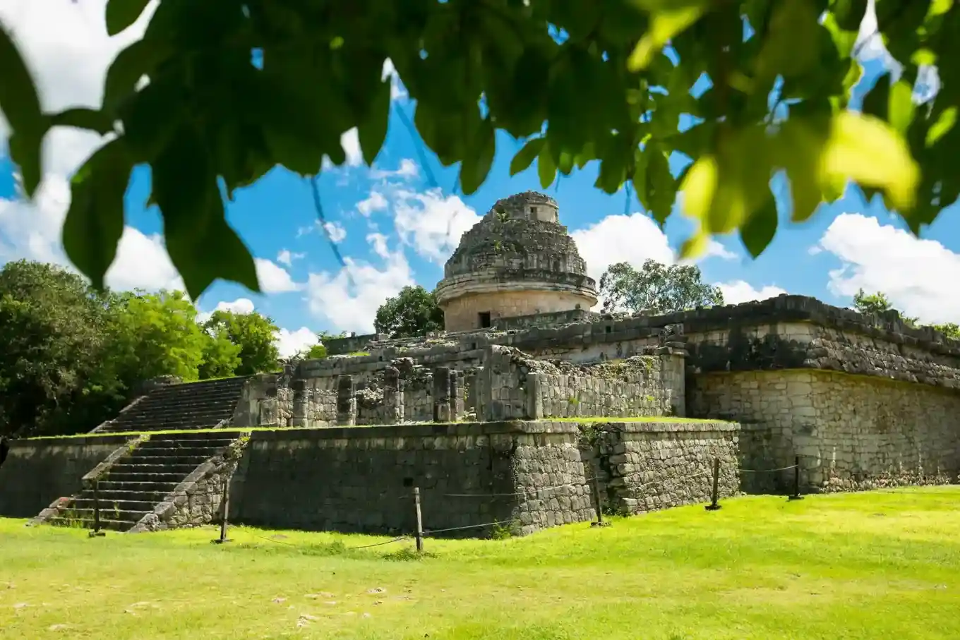 Ancient El Caracol observatory at Chichen Itza ruins framed by green foliage and a bright blue sky.
