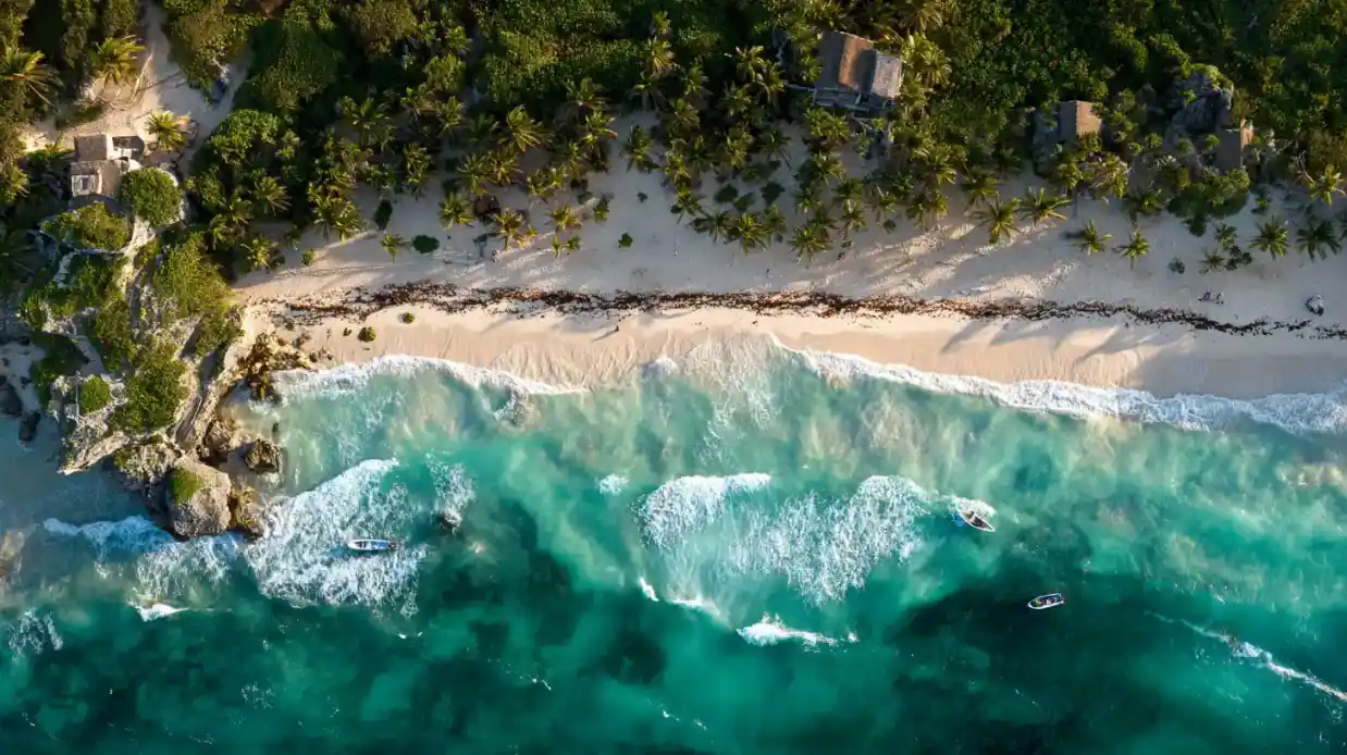 Aerial view of Tulum beach with turquoise waves, palm trees, and boats at sunset.