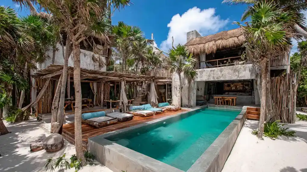 Luxury beachfront villa in Tulum with private pool, wooden lounge chairs, and palm trees.